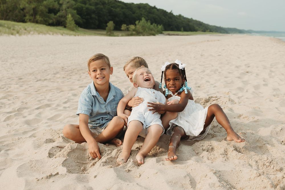 Four young children sitting together on a sandy beach, smiling and laughing on a sunny day.