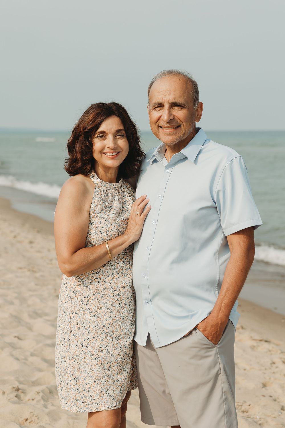 Smiling middle-aged couple posing together on a sandy beach with ocean waves in the background.