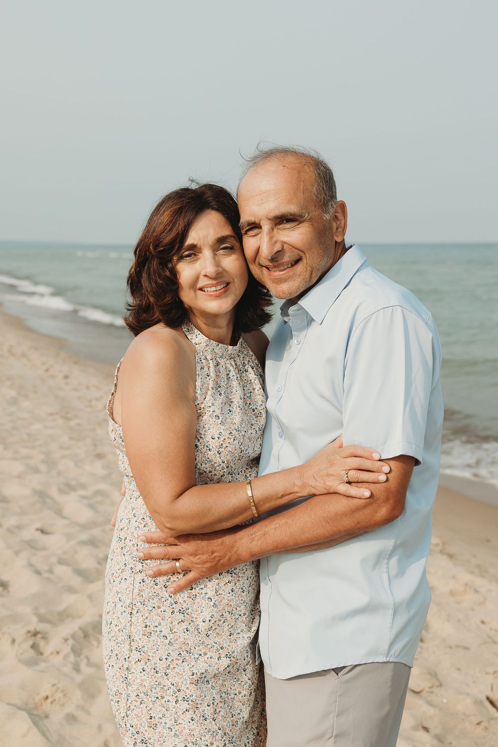 A smiling middle-aged couple embracing on a sandy beach with ocean waves in the background.