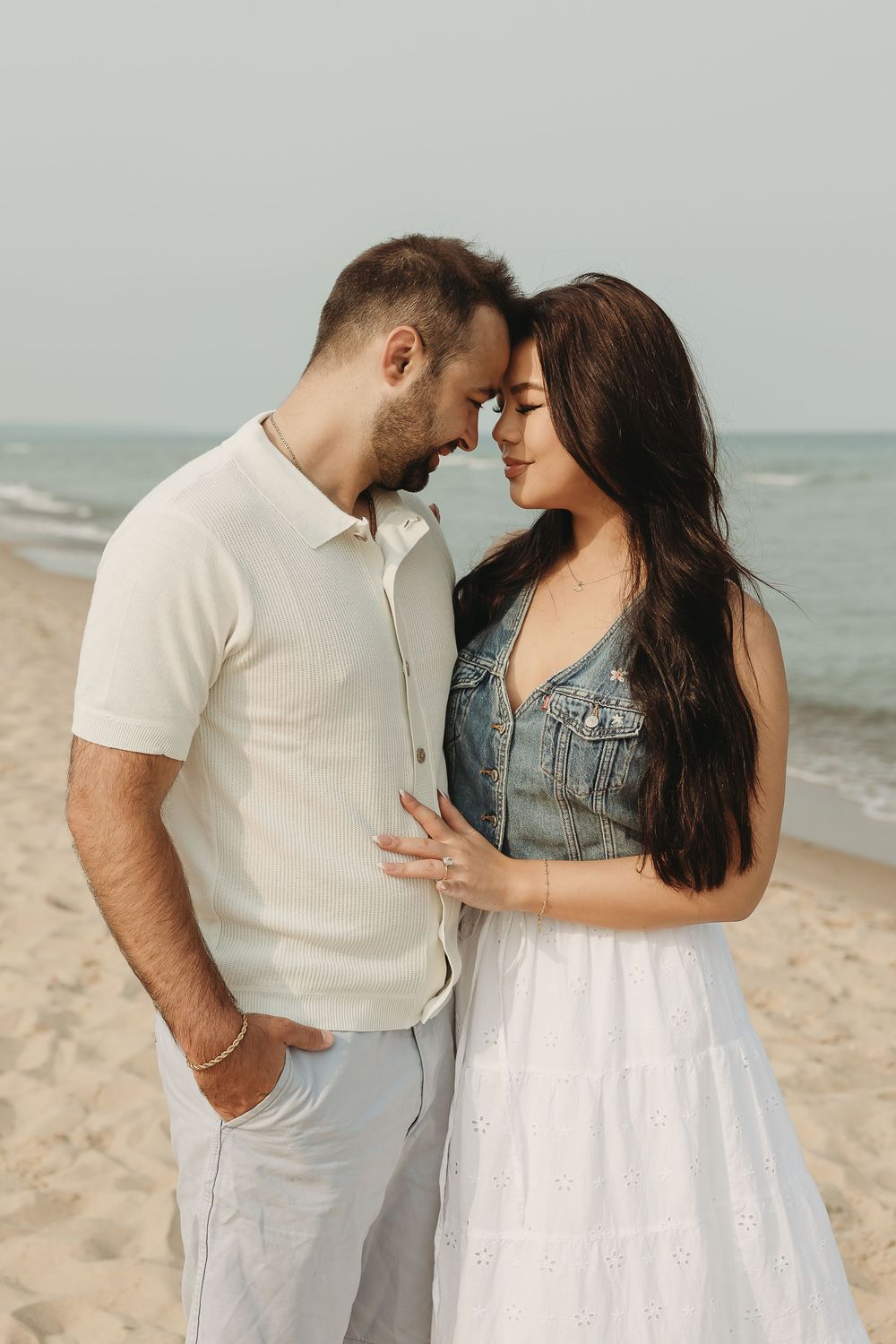 Romantic couple touching foreheads on a sandy beach, woman in denim vest and white skirt, man in cream shirt.