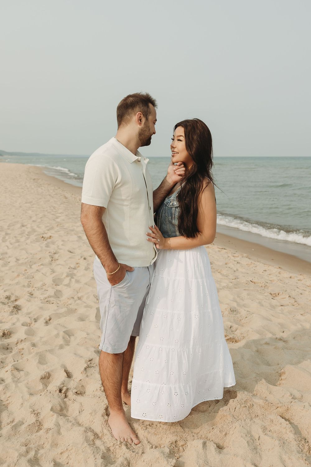 A couple embraces on a sandy beach, the woman in a white dress and man in white shirt and shorts, with ocean waves behind them.
