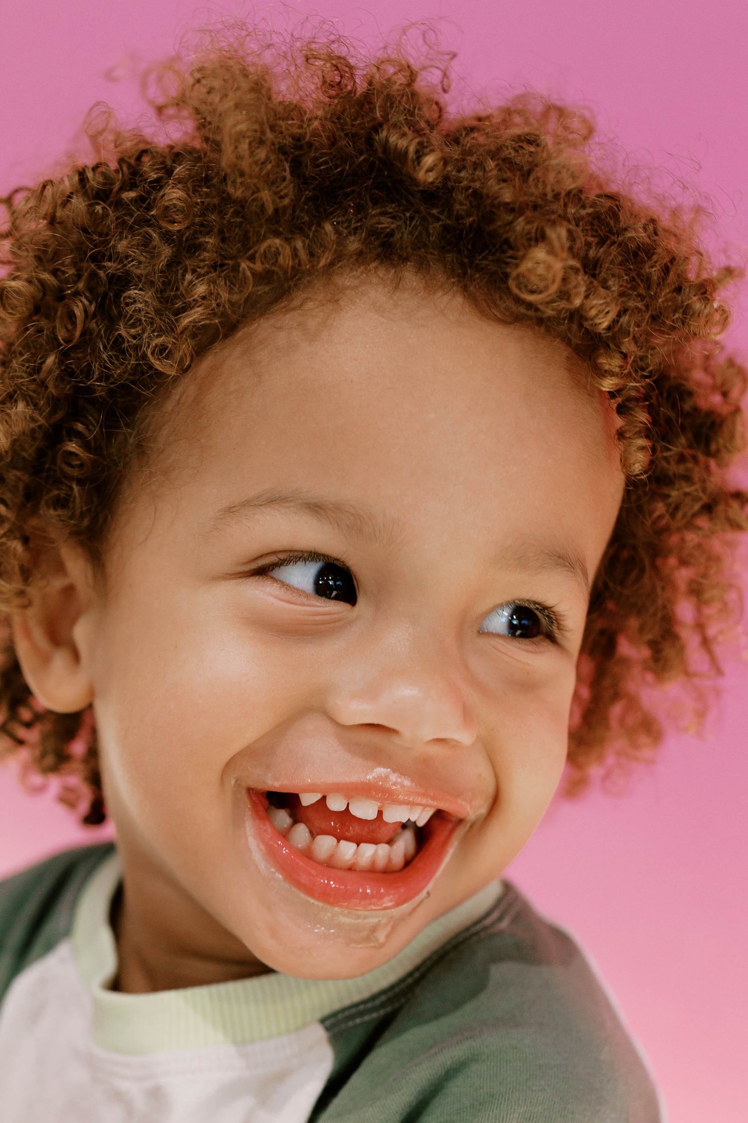Close-up portrait of a laughing toddler with curly hair against a bright pink background.