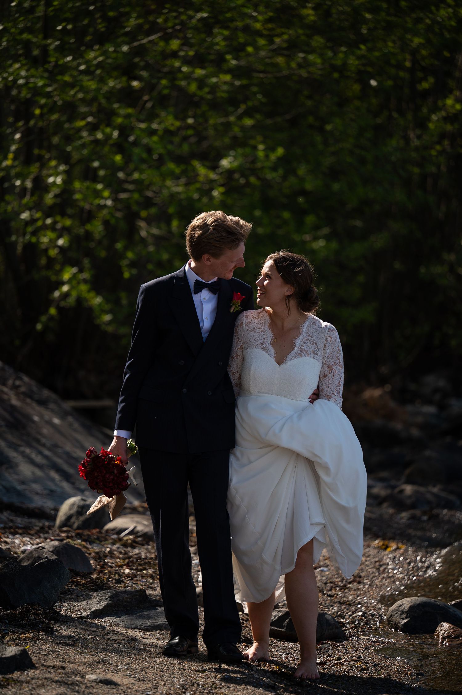 Bride in lace gown and groom in tuxedo share romantic moment on rocky outdoor setting with lush green trees.