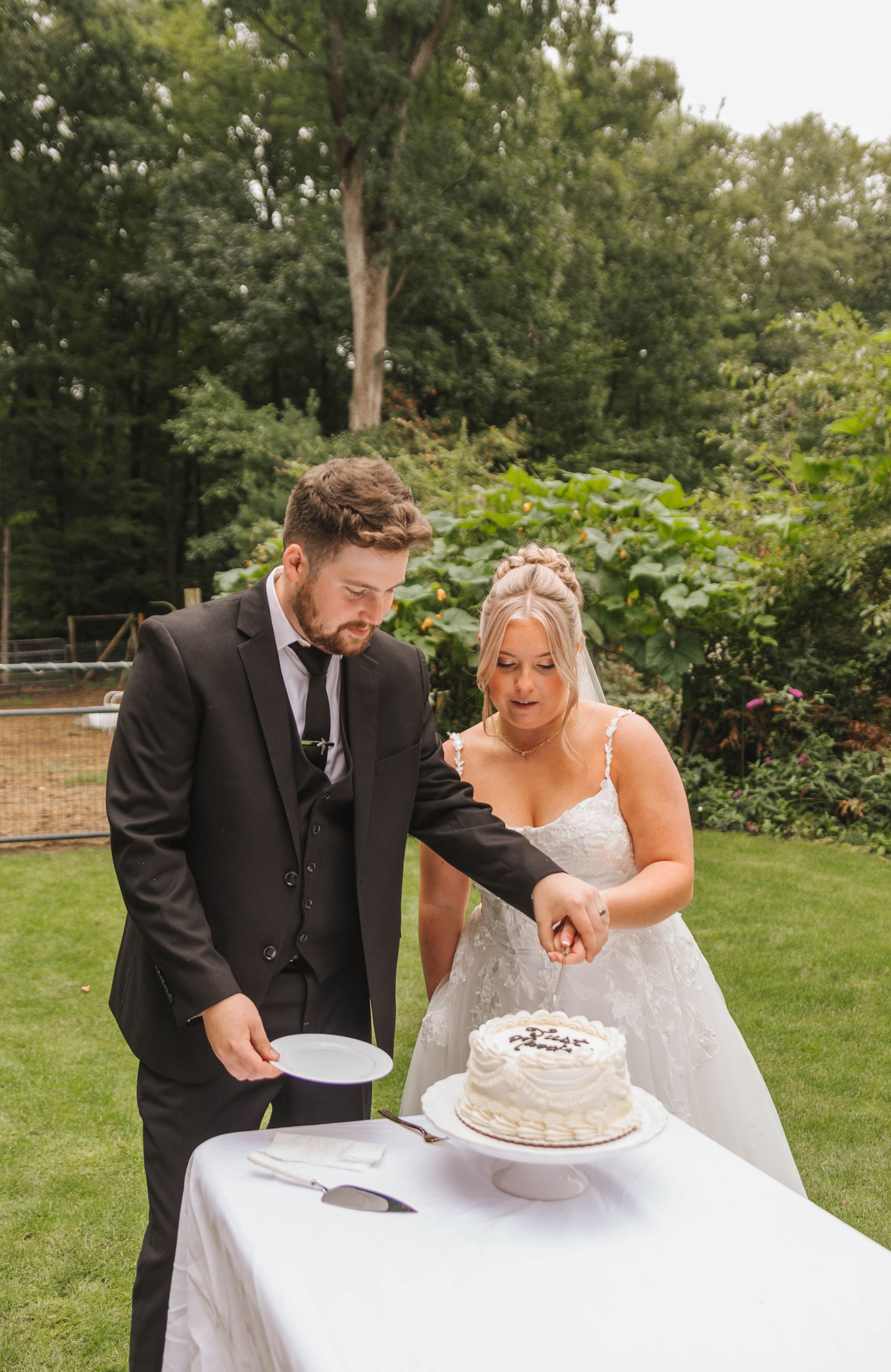 Bride and groom cutting their white wedding cake together outdoors on a sunny day.