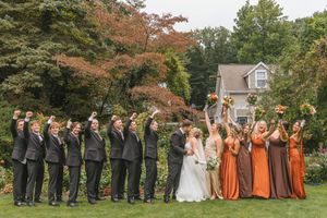 Wedding party in autumn garden, groomsmen in gray suits and bridesmaids in burnt orange dresses cheering with bouquets raised.