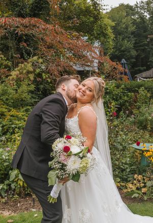 Bride in white lace gown holds floral bouquet as groom in gray suit kisses her cheek in a lush garden setting.