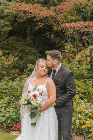 Groom kisses bride on cheek as she smiles holding red and white bouquet in lush garden wedding portrait.