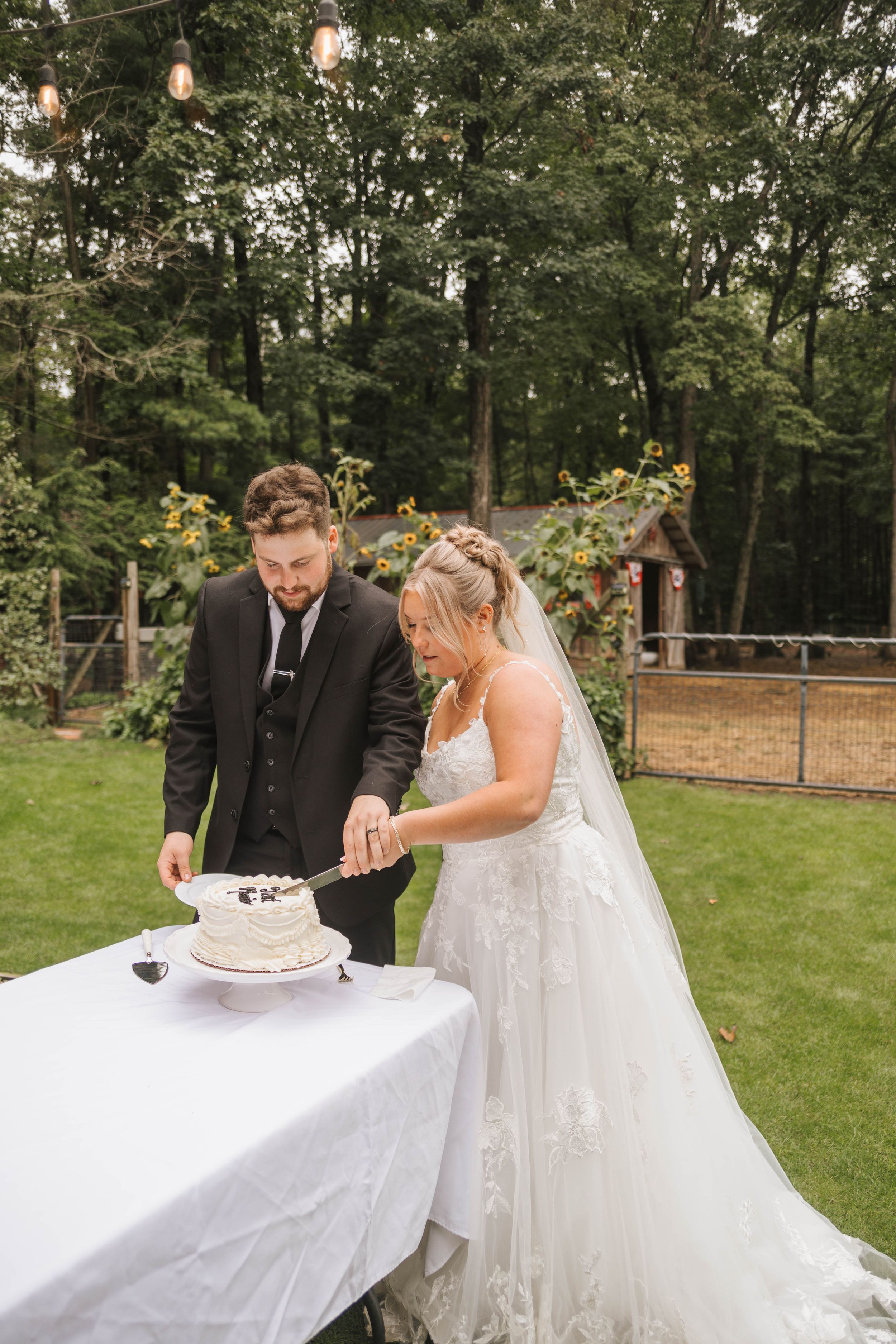 Bride and groom cutting their white wedding cake outdoors on a lace-covered table surrounded by lush green trees.
