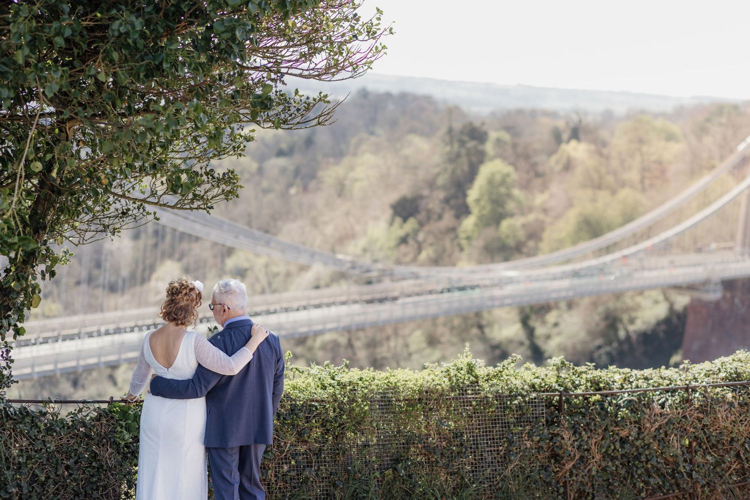 Bride and groom embracing outdoors with a suspension bridge and lush green valley in the background on their wedding day.
