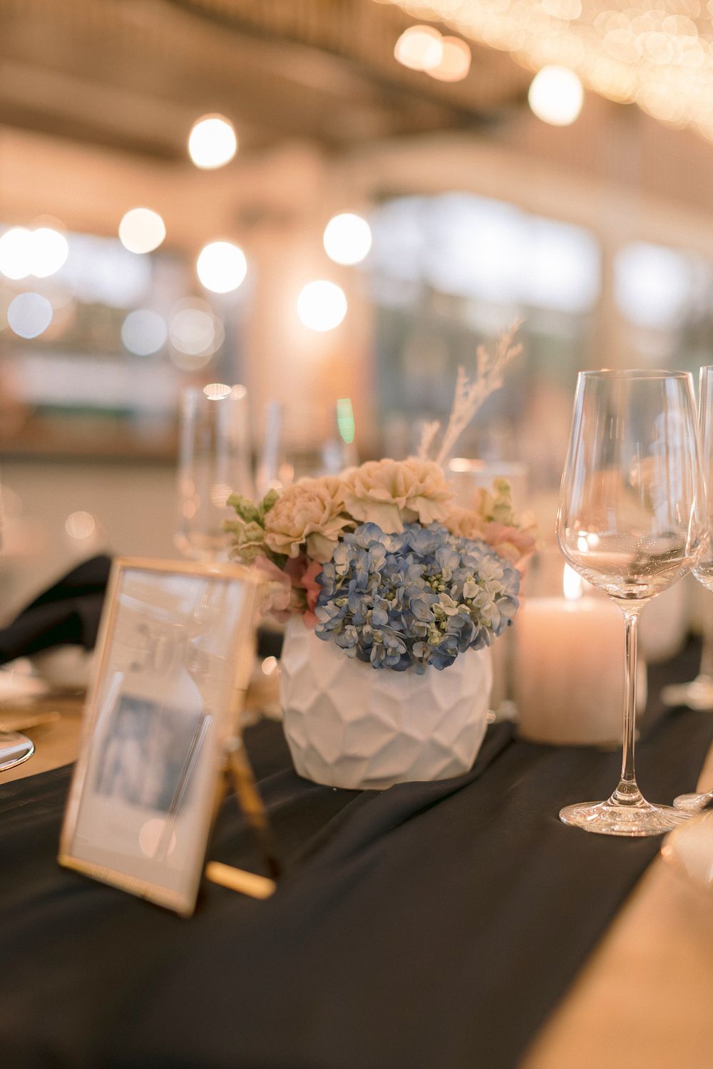 Elegant wedding reception table with blue hydrangeas in white geometric vase, champagne flute, candle, and framed photo.