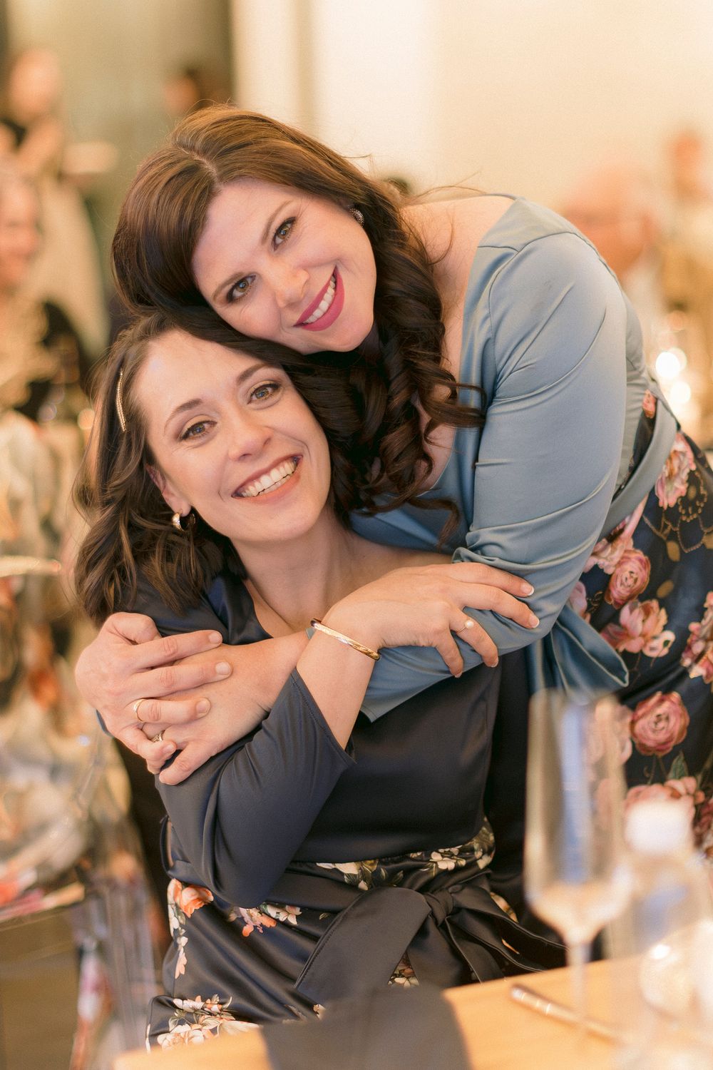 Two smiling brunette women embrace warmly at an elegant event, one leaning over the other's shoulder.