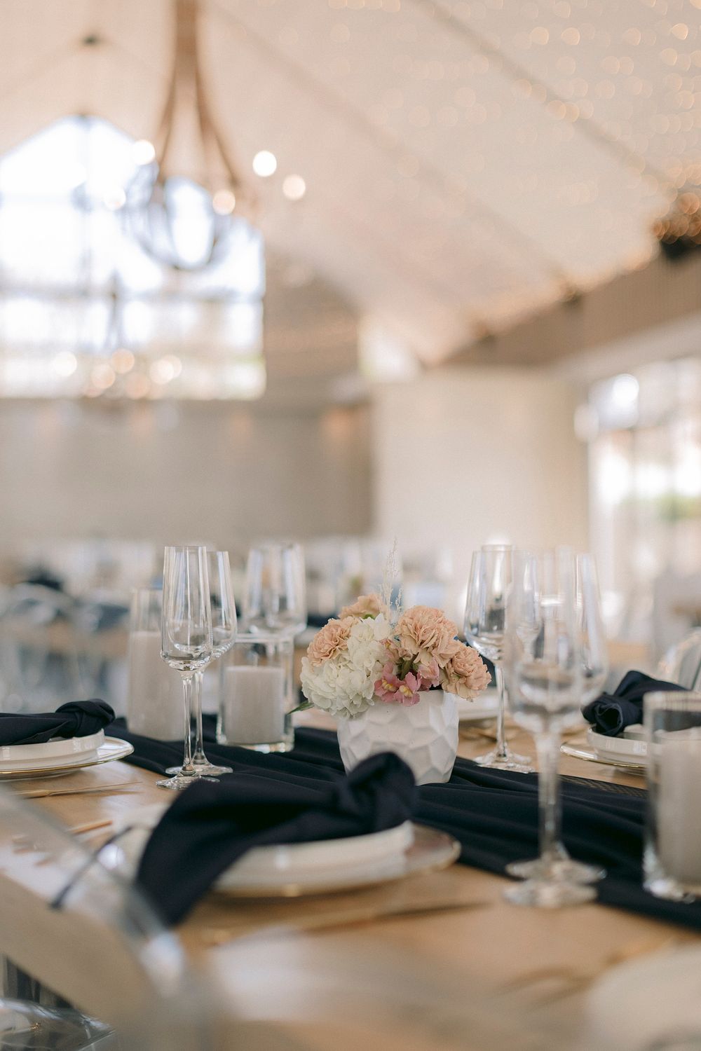 Elegant wedding reception table set with navy napkins, crystal glasses, and a blush floral centerpiece in a bright venue.