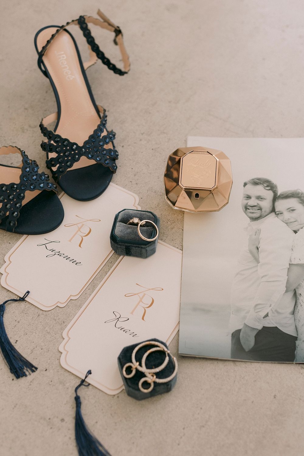 Elegant wedding flat lay with navy heels, gold ring box, wedding bands, monogram cards, and a vintage couple photo on neutral background.