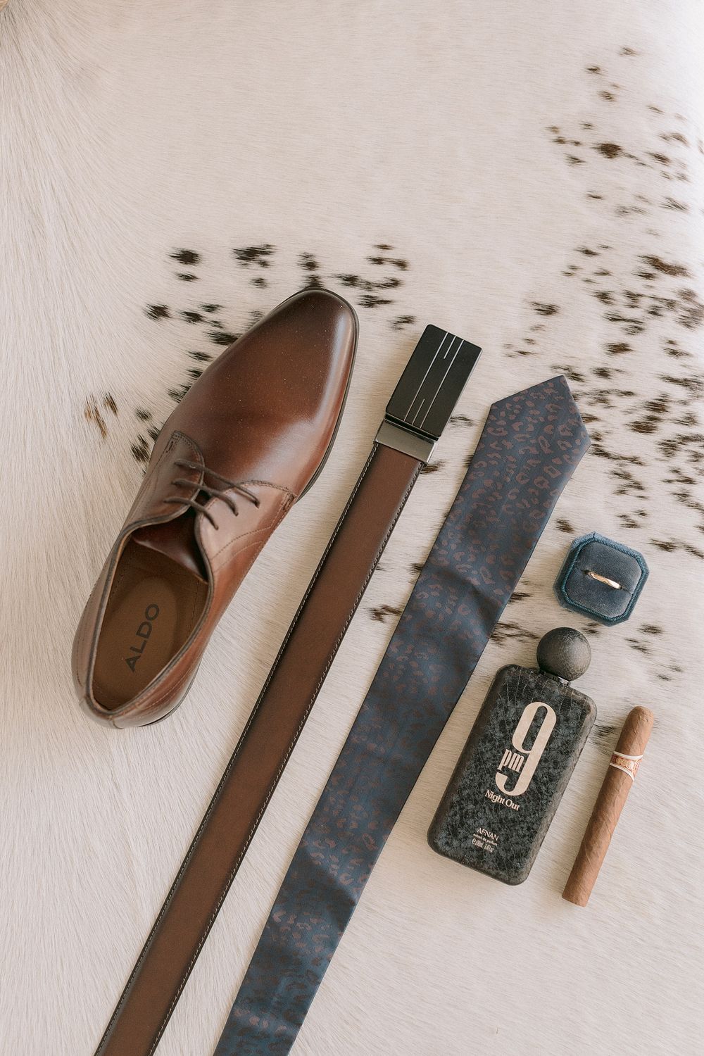 Brown leather Oxford shoe, belt, blue tie, cologne, cigar, and ring box arranged as groom wedding day accessories.