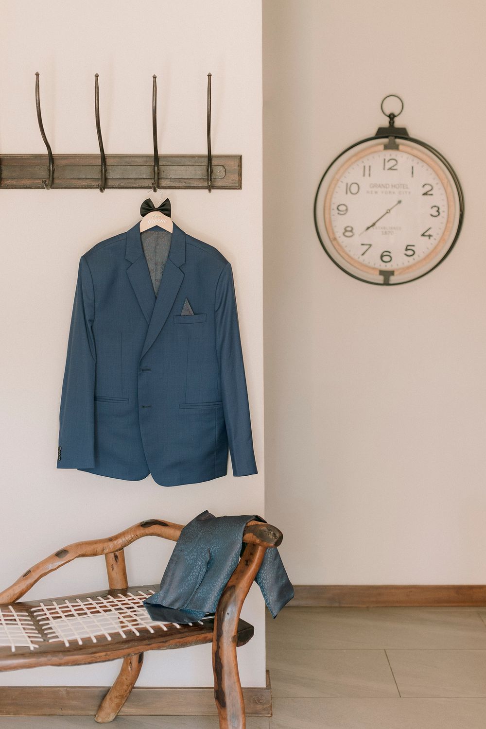 Blue suit jacket hanging on wall hooks with bow tie, wooden bench with trousers, and vintage wall clock in elegant room.