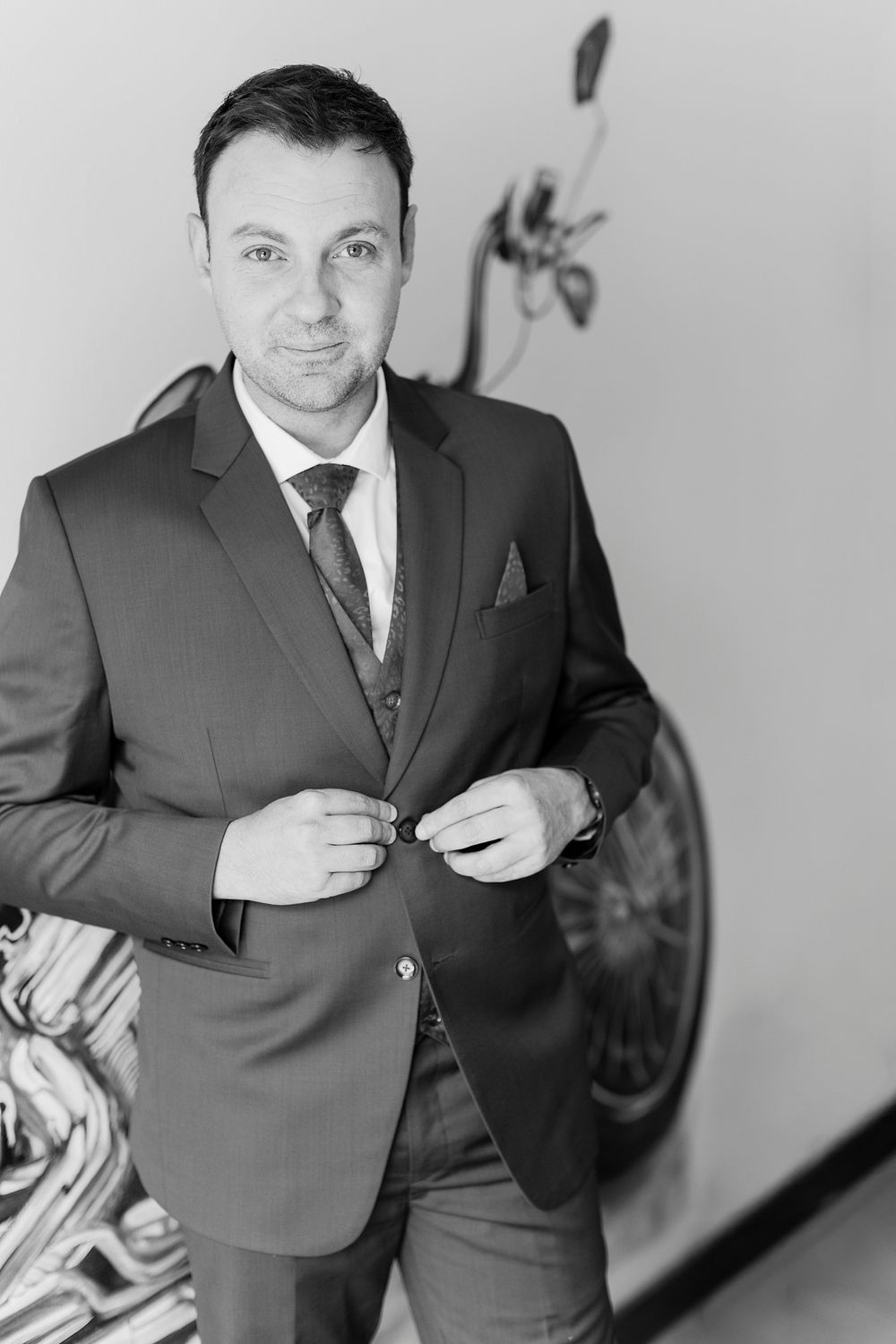 Black and white portrait of a well-dressed man in a suit adjusting his jacket button, wearing a tie and pocket square.