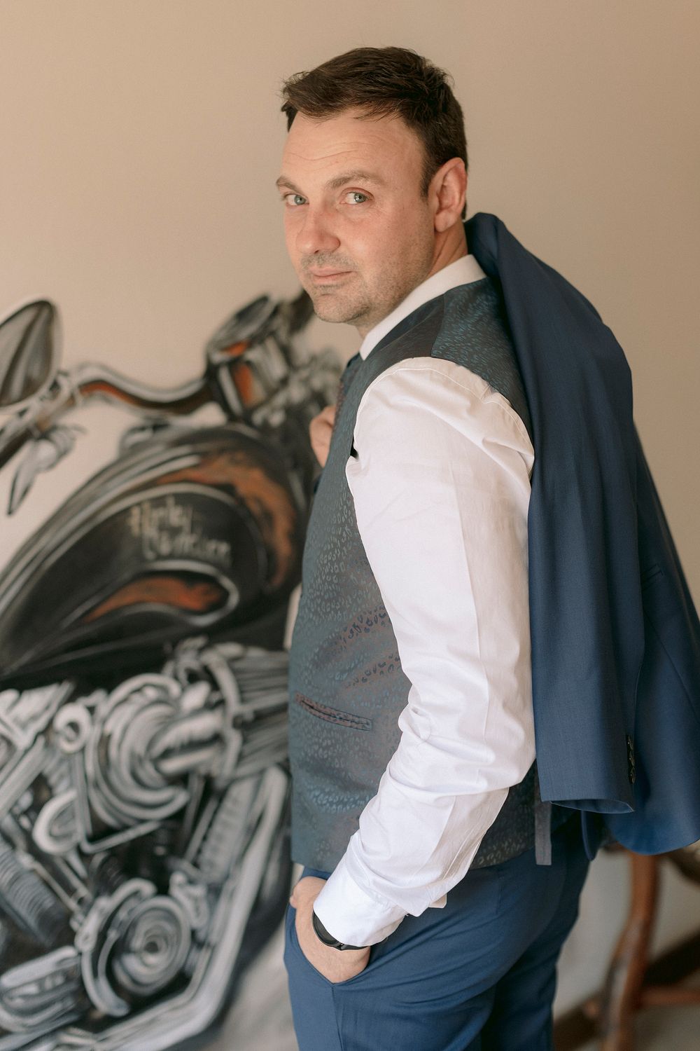 A well-dressed man in a vest and white shirt holds a blue blazer over his shoulder near a motorcycle mural.