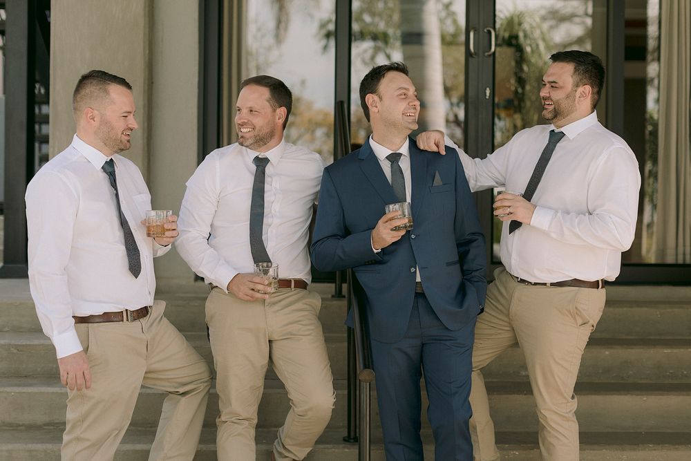 Groom in navy suit laughs with three groomsmen in white shirts and ties holding drinks outside a venue.