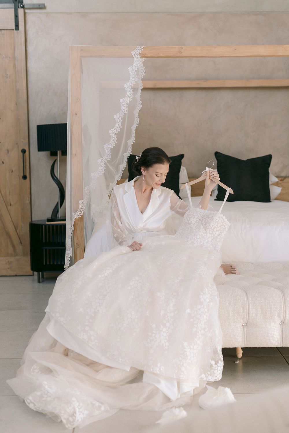 Bride in white robe sits on bed holding lace wedding dress on hanger, with veil and elegant bedroom decor.