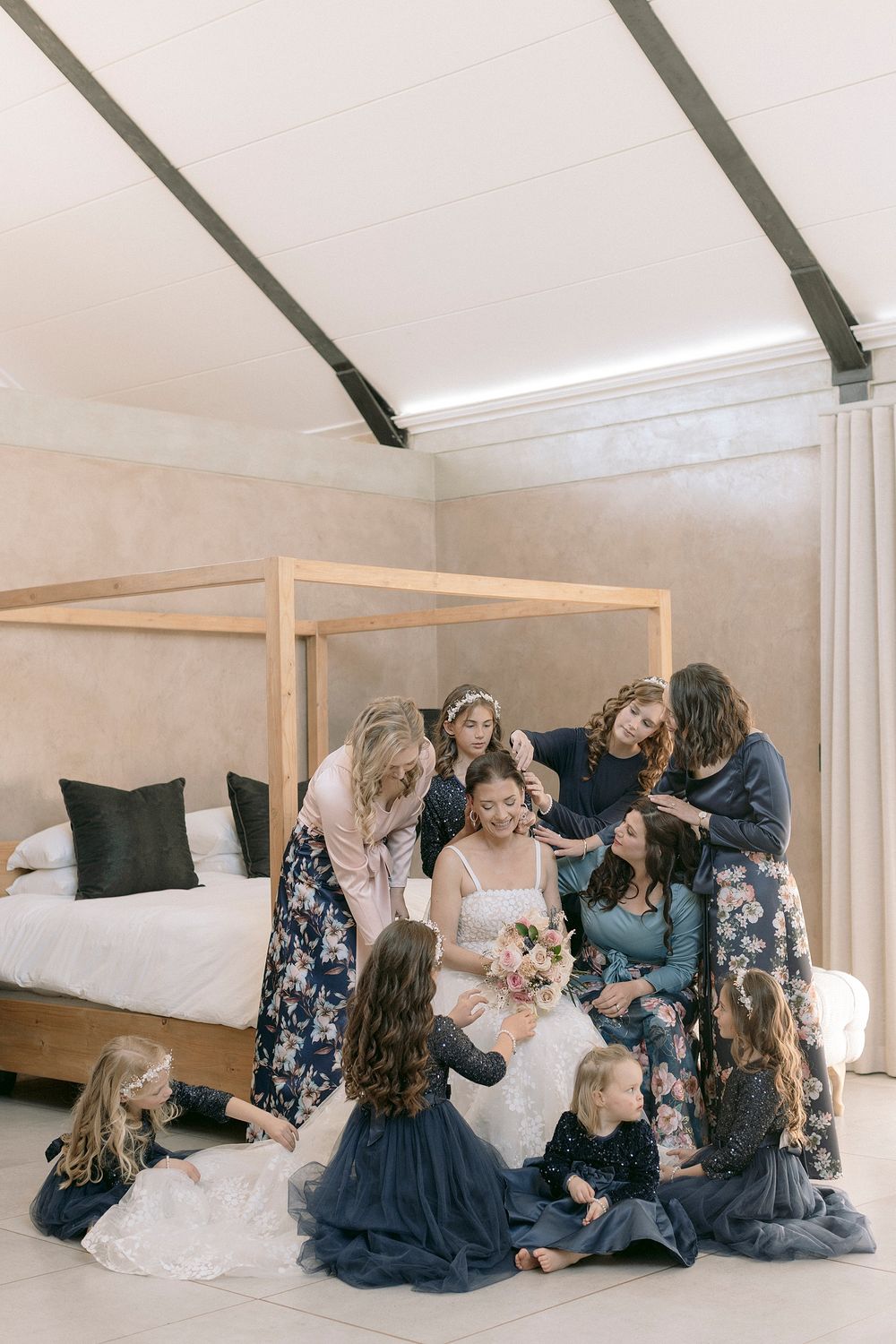 Bride in white gown surrounded by bridesmaids in navy dresses during wedding preparations in elegant room.