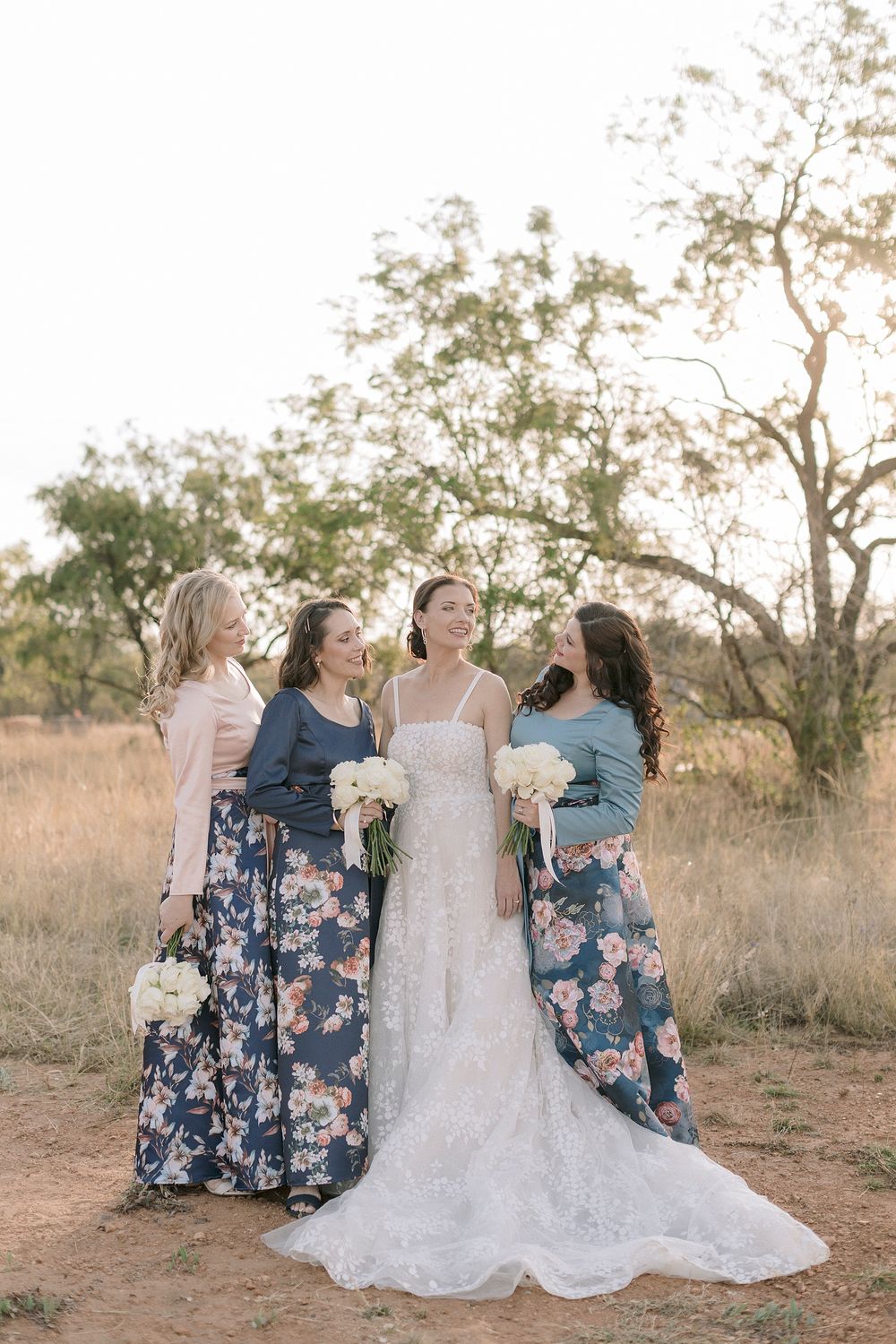 Bride in white lace gown with three bridesmaids in floral skirts holding bouquets outdoors at golden hour.