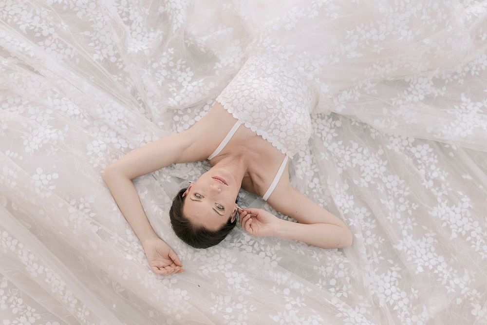 A bride in a white lace gown lies gracefully on flowing embroidered fabric, gazing upward in a serene bridal portrait.