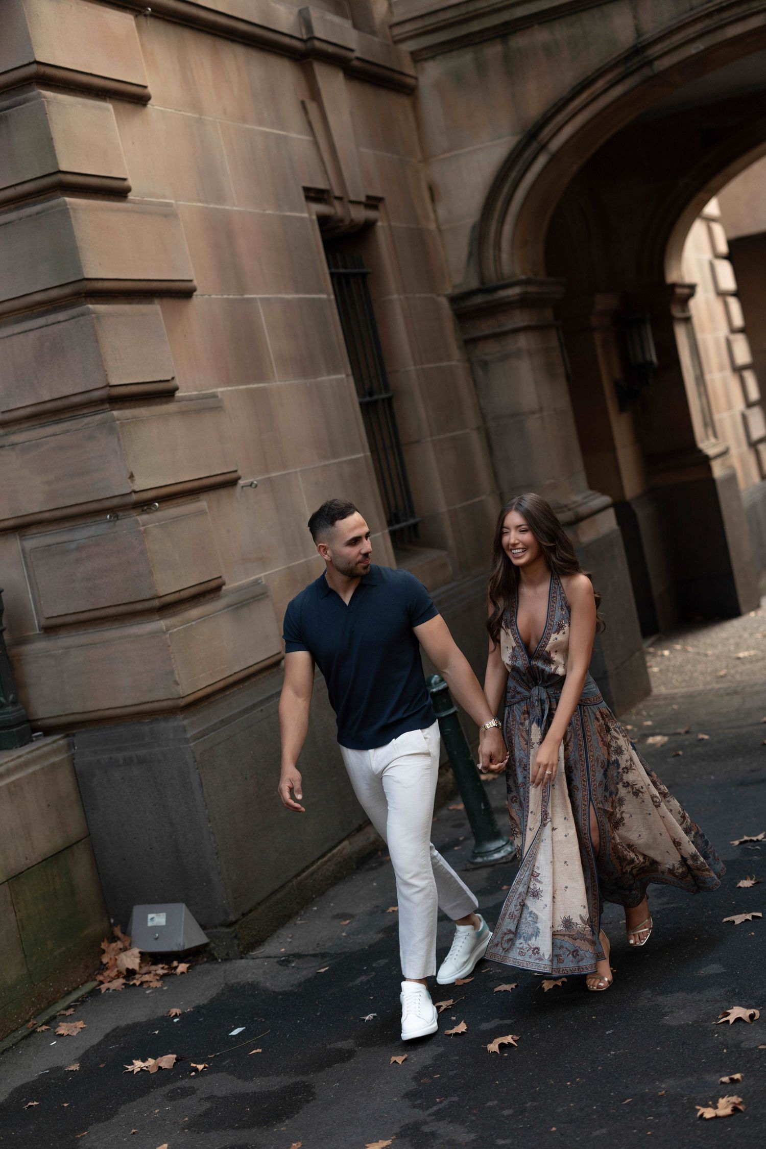 A couple holding hands and walking together past historic stone architecture, woman in floral maxi dress, man in navy shirt and white pants.