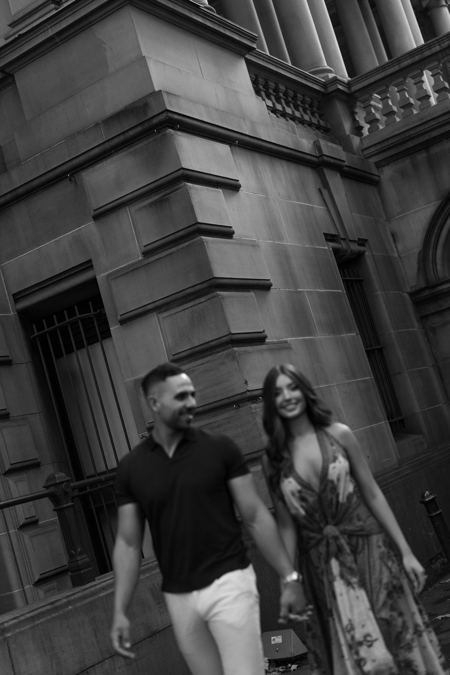 Black and white photo of a stylish couple holding hands and smiling while walking past a classic stone building.