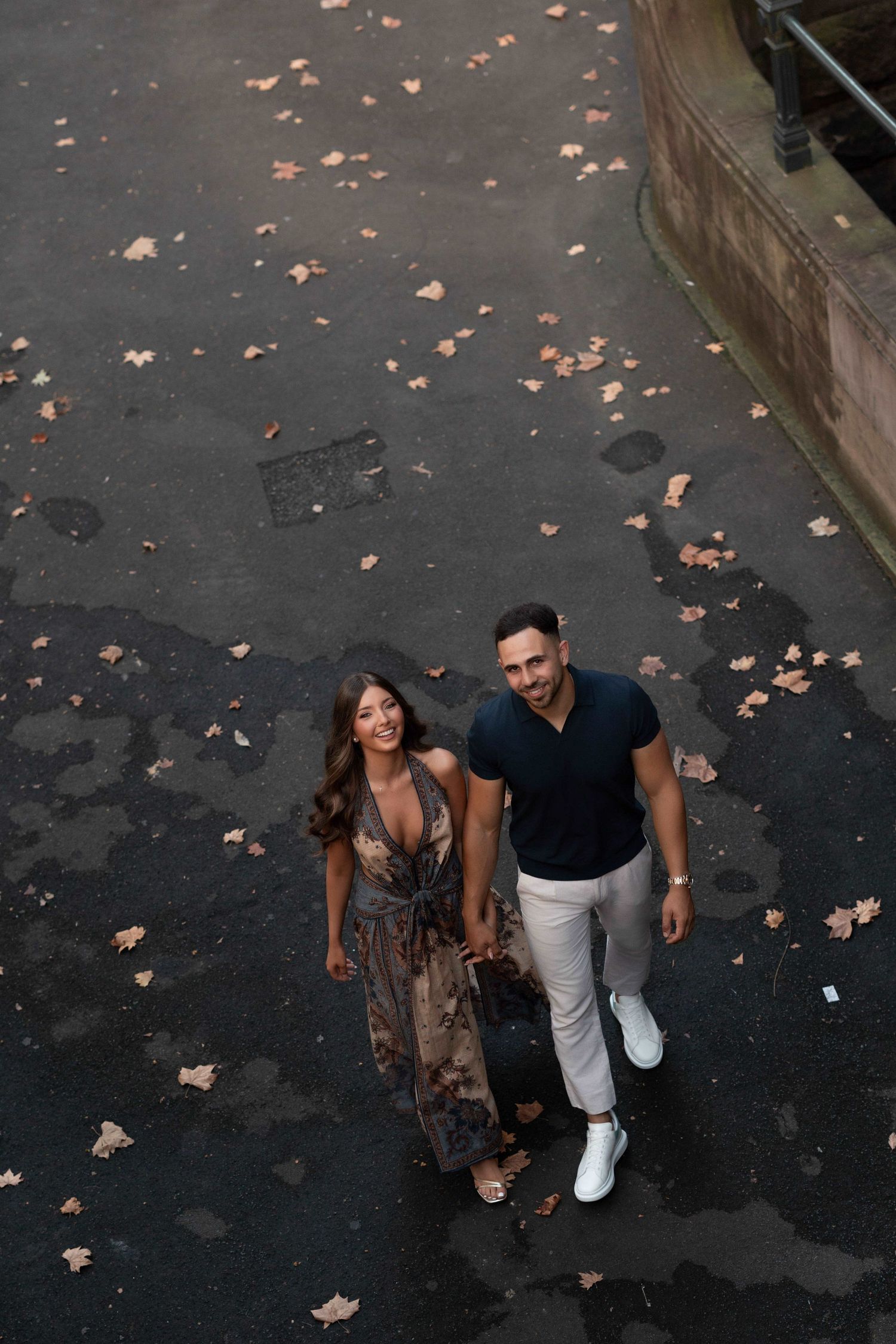 Couple holding hands walking on autumn leaf-covered pavement, woman in floral dress and man in dark shirt and white pants.
