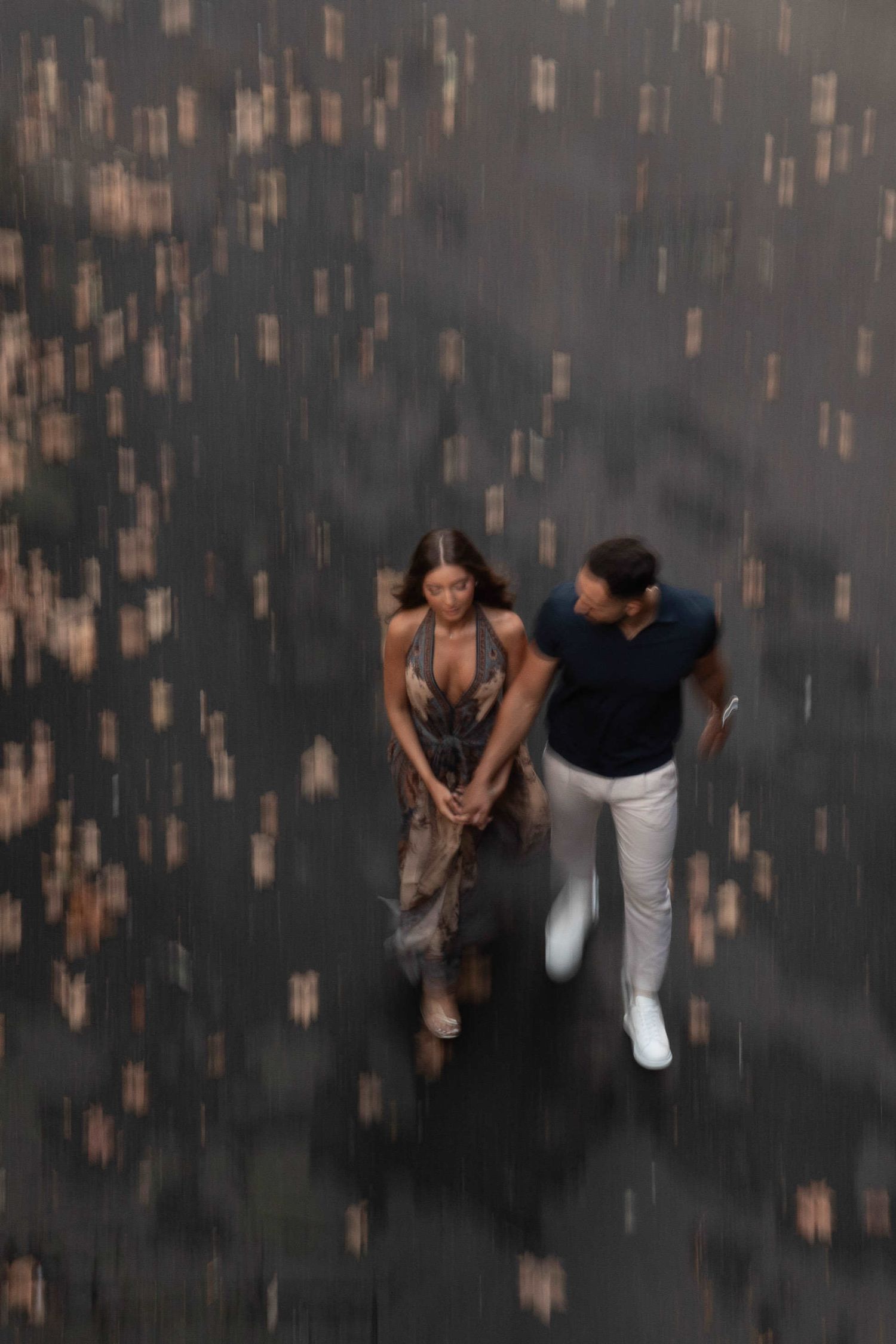 Aerial view of a couple holding hands walking in the rain, woman in floral dress, man in dark shirt and white pants.