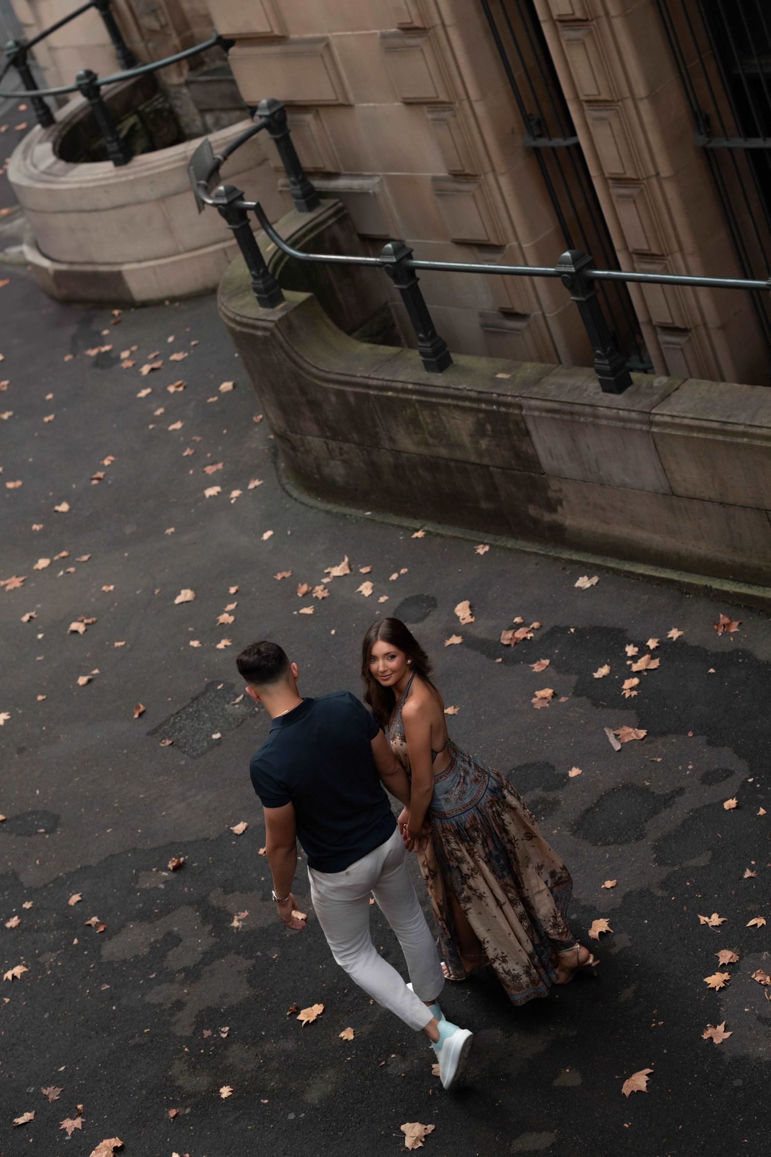 Aerial view of a couple holding hands walking on a city street with autumn leaves scattered on the ground.