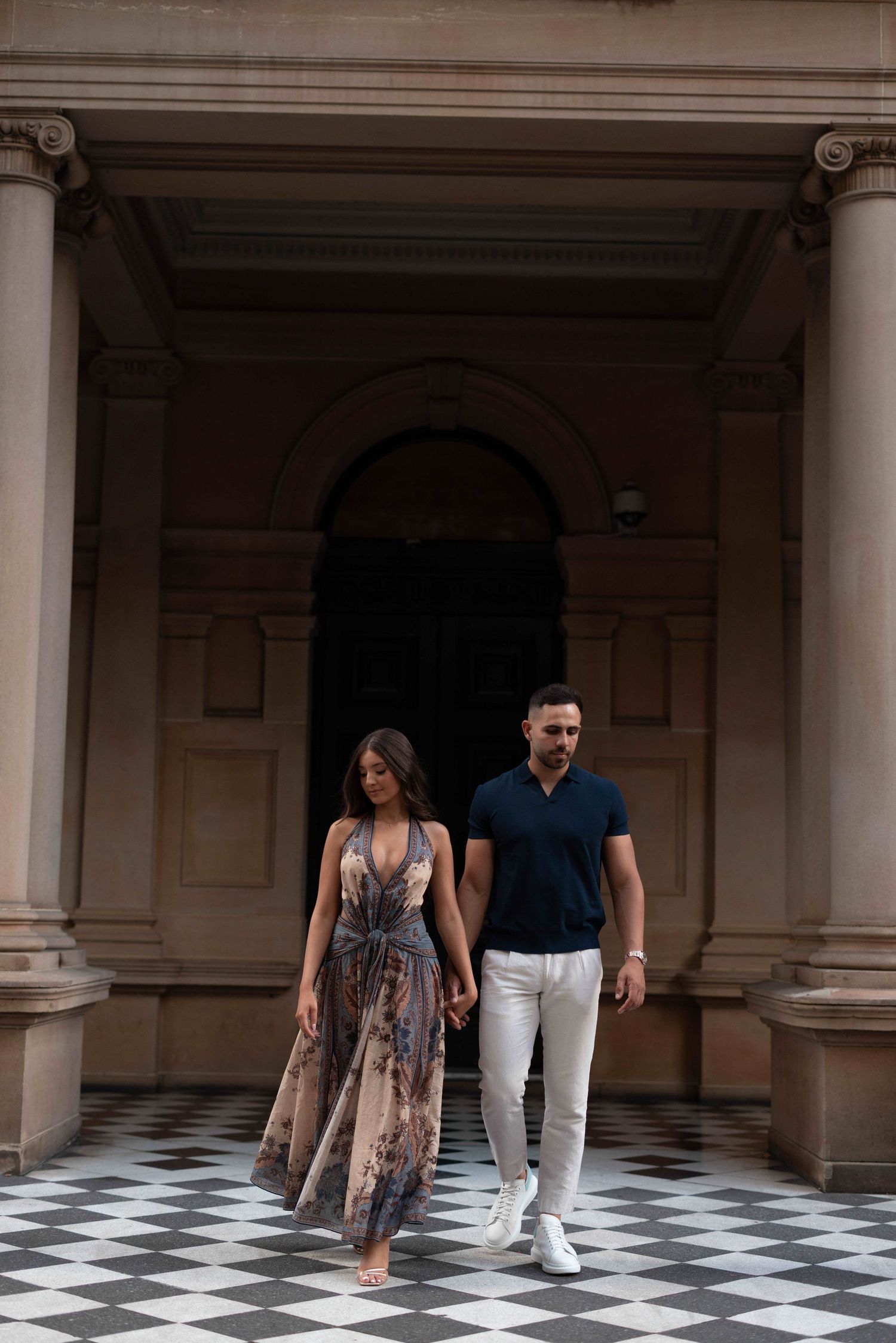 Couple walking hand in hand on checkered floor outside elegant stone building with columns.
