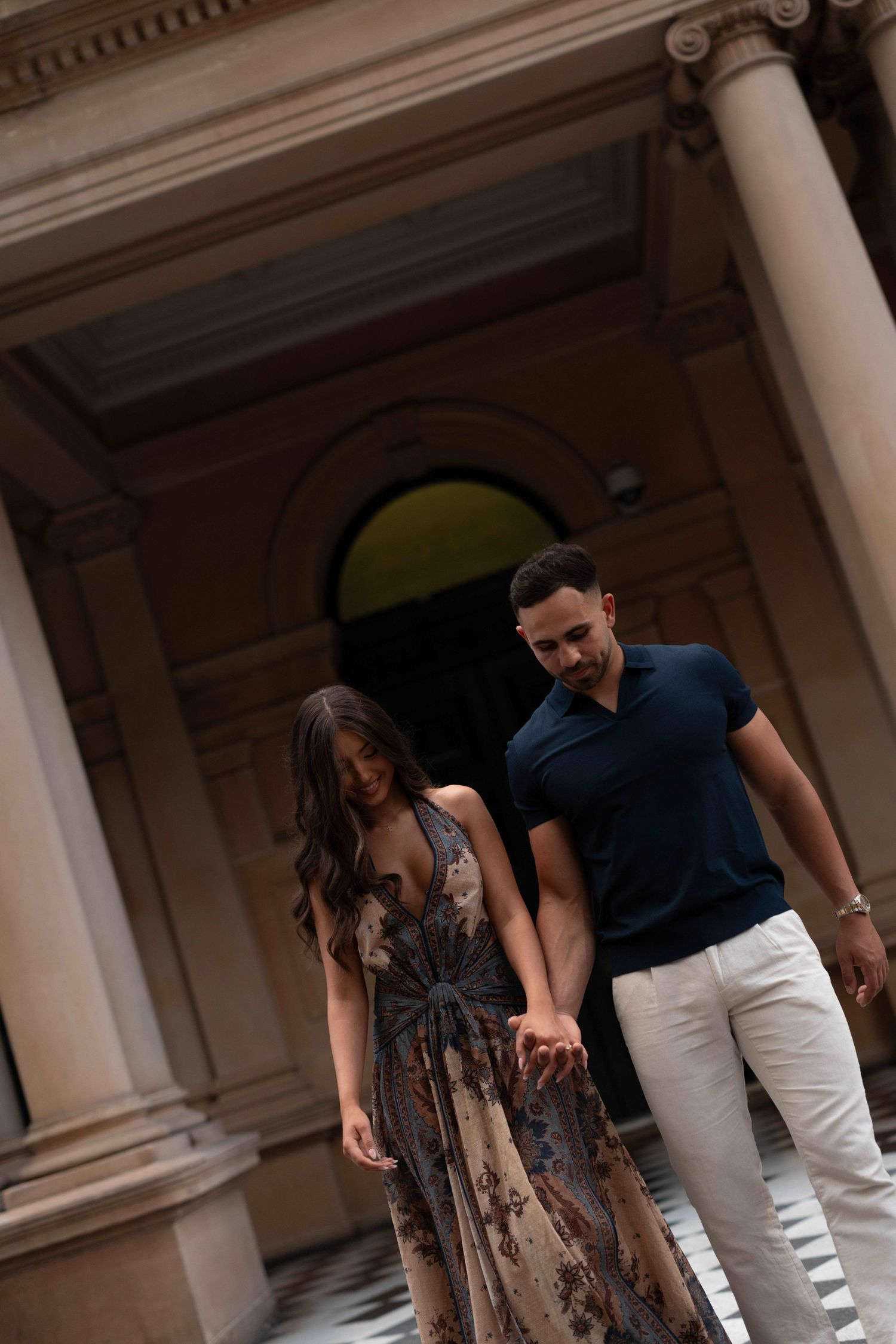 Couple holding hands in front of elegant building with columns, woman in floral maxi dress, man in navy shirt and white pants.