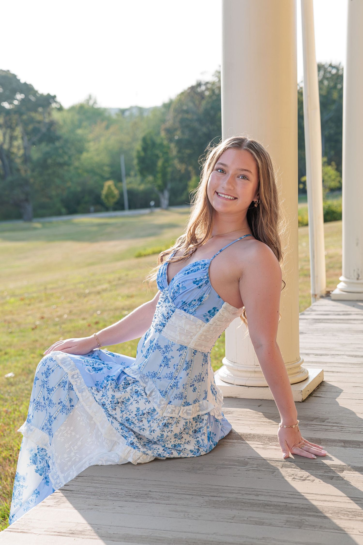 Smiling young woman in blue floral sundress sits on wooden porch with white columns in sunlight. Wells Reserve at Laudholm Wells ME