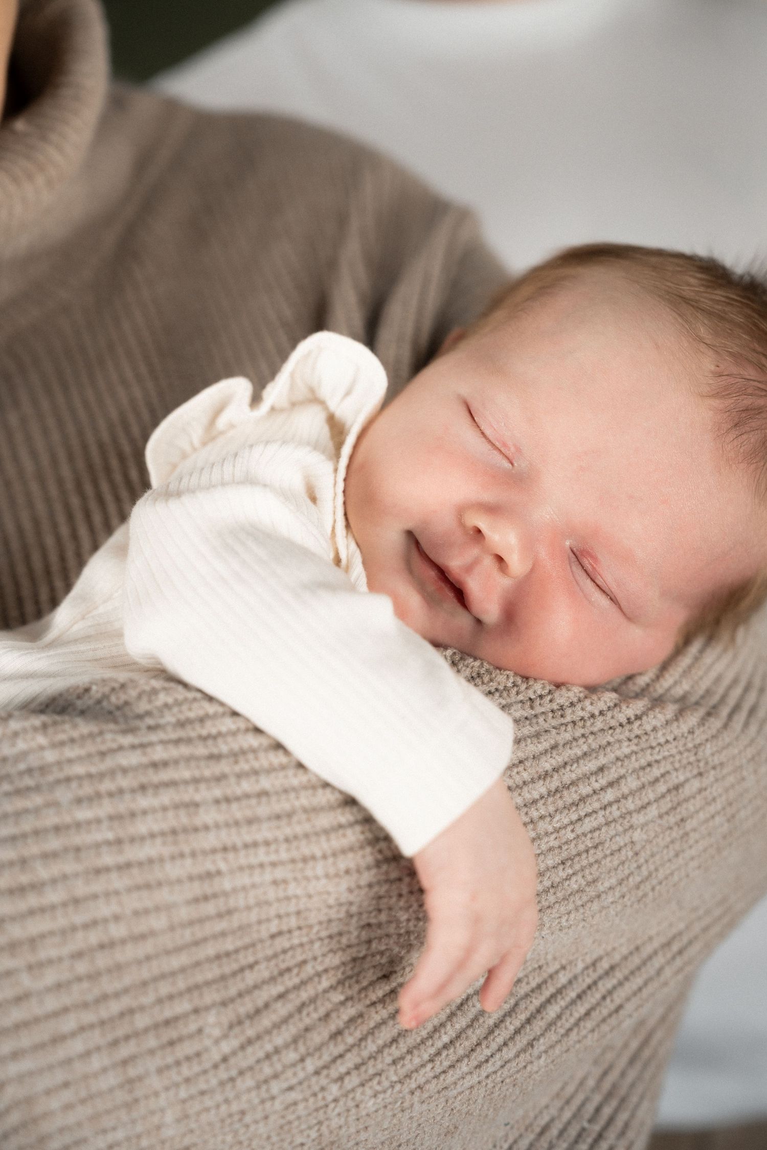 Sleeping newborn baby in white onesie resting peacefully in the arms of a caregiver wearing a beige knit sweater.