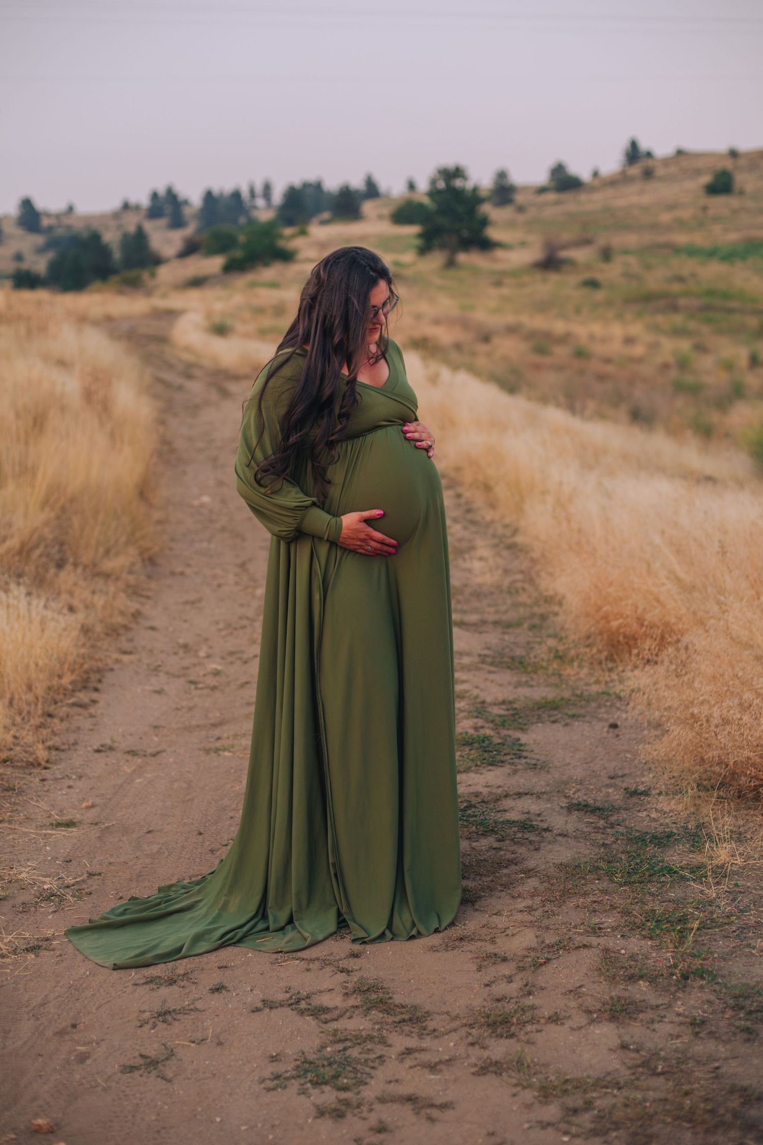 Pregnant woman in elegant olive green maternity gown standing on a dirt path in a golden field.