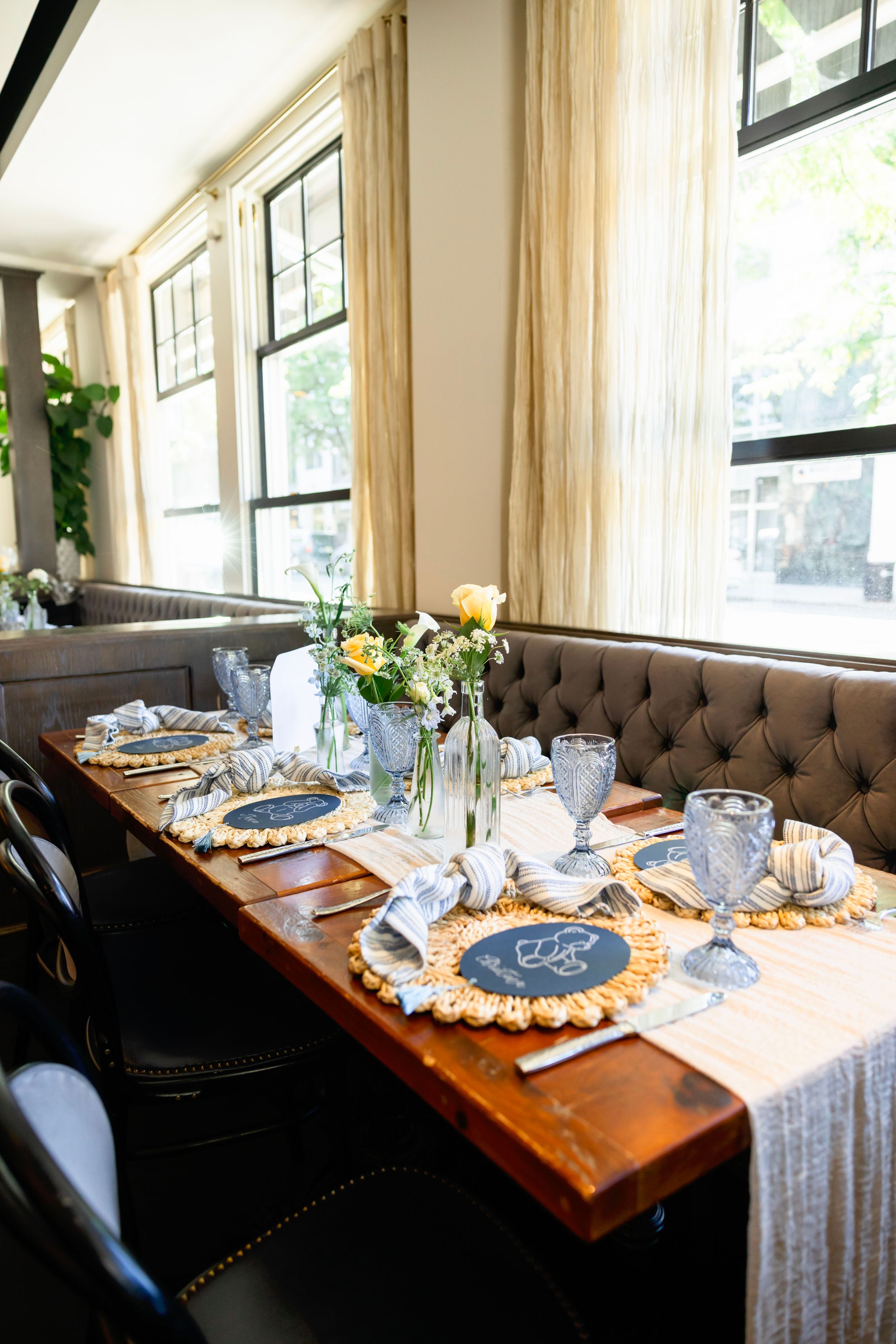 Elegant restaurant dining table with chalkboard chargers, blue goblets, gold woven placemats, and fresh floral centerpiece by tufted booth.