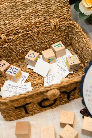 Wooden alphabet blocks and handwritten cards inside a woven wicker basket labeled TOYS at a baby shower.