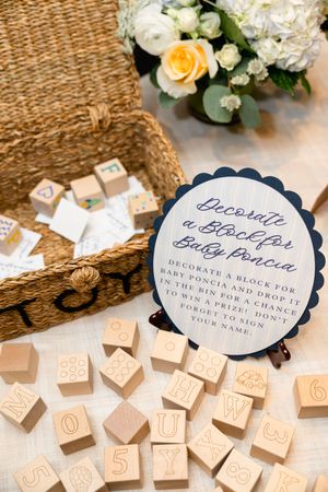 Baby shower block decorating station with wooden alphabet blocks, wicker basket, and floral arrangement on white tablecloth.