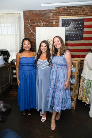 Three women in blue dresses smile together indoors near an American flag and brick wall backdrop.