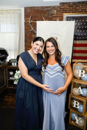 Two women pose together at a baby shower, one pregnant in a blue dress, with BABY letter boxes and American flag decor.