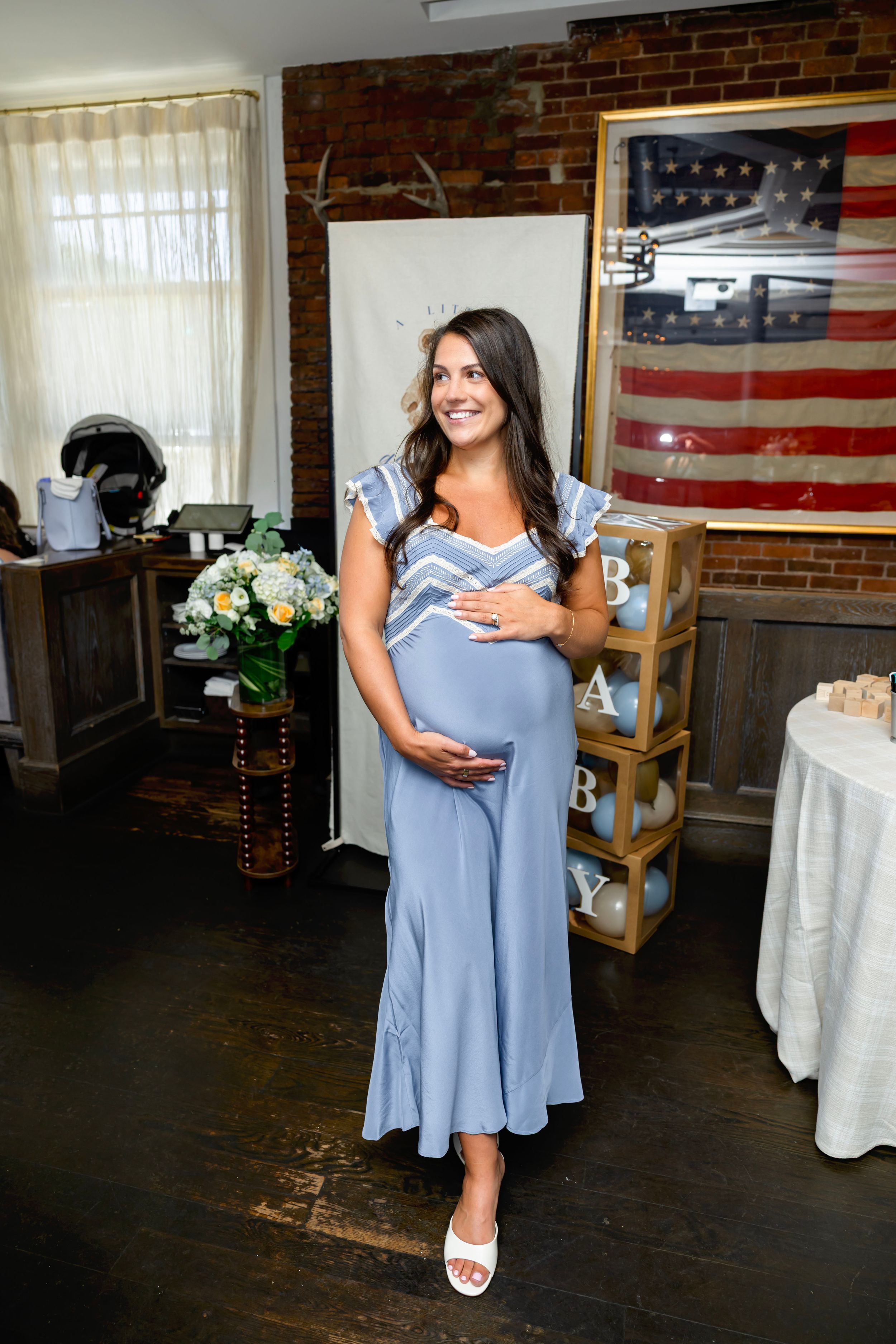 Pregnant woman in blue midi dress holding baby bump at a baby shower with American flag decor.