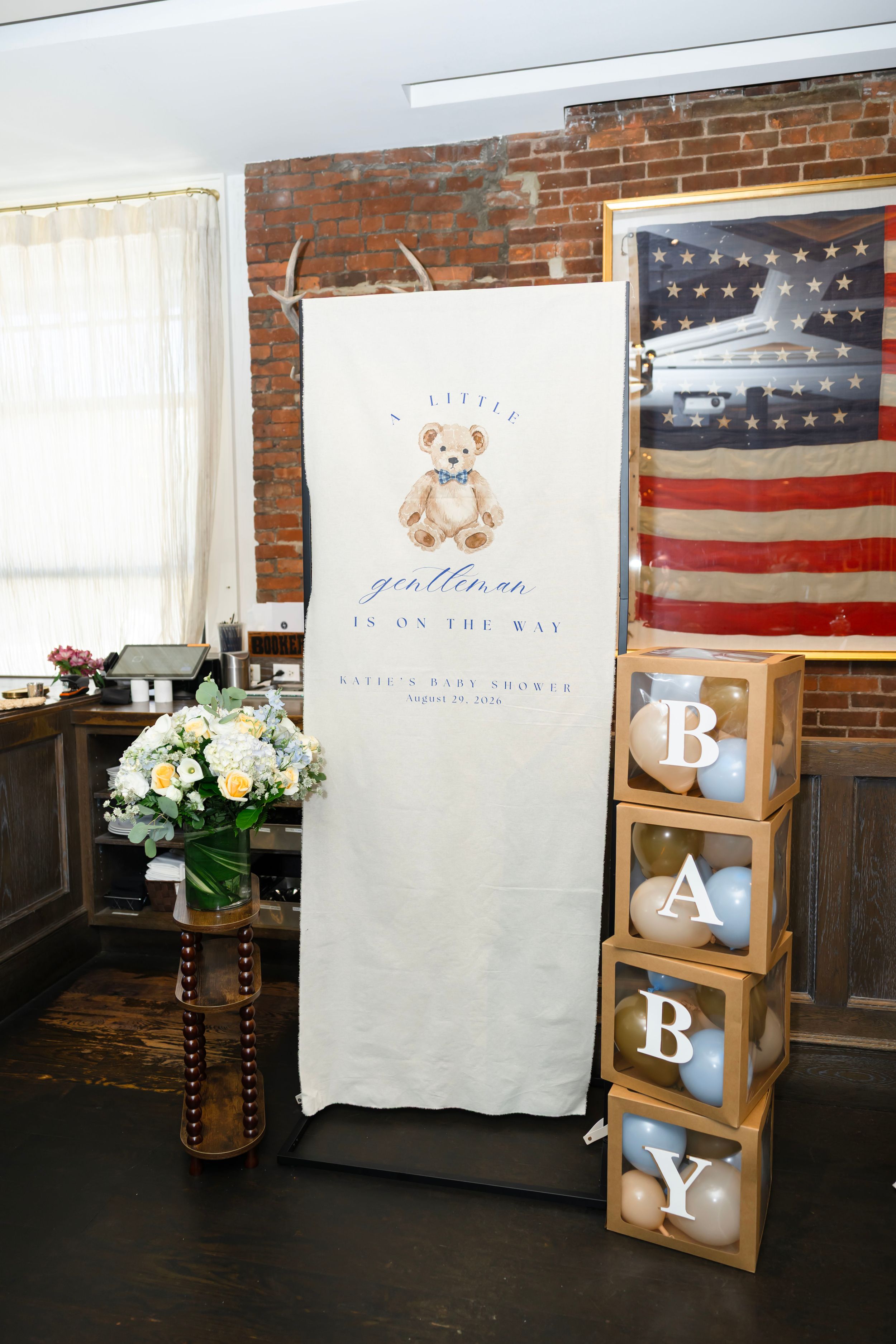 Baby shower decor with teddy bear banner, BABY letter blocks, floral arrangement, and American flag backdrop.