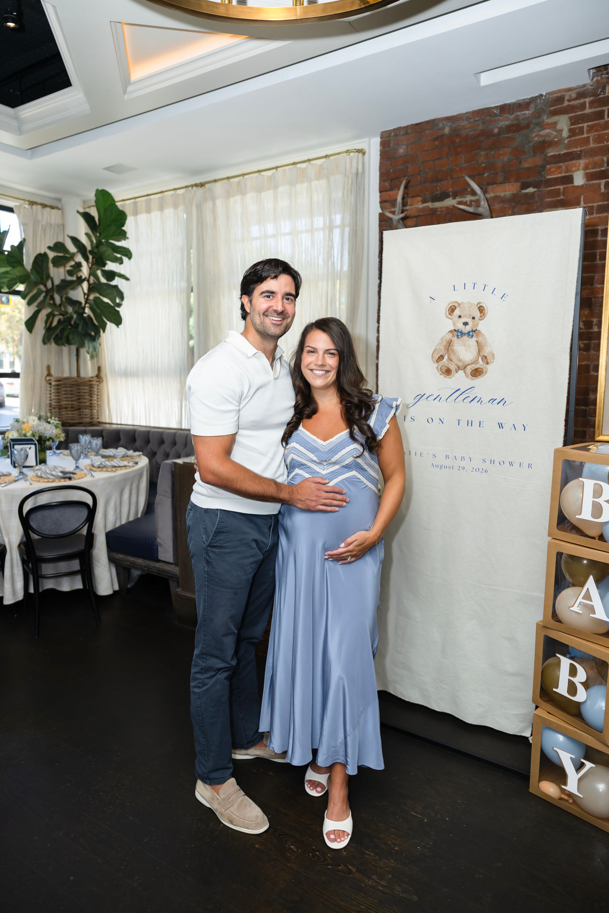Couple poses at baby shower with "Little Gentleman on the Way" banner, pregnant woman in blue dress holding belly.
