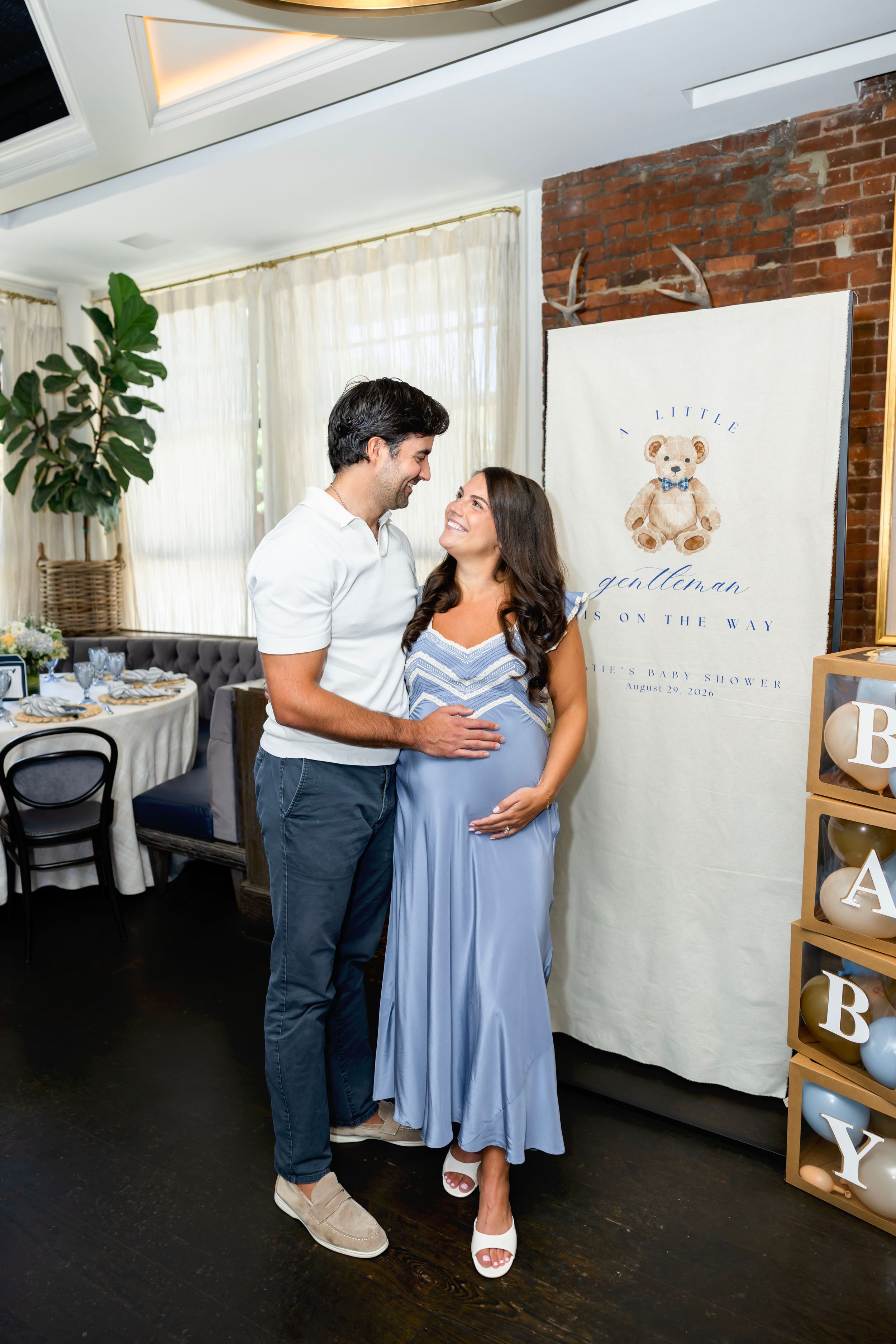 Couple at baby shower, woman in blue dress cradling baby bump, standing before a teddy bear themed backdrop.