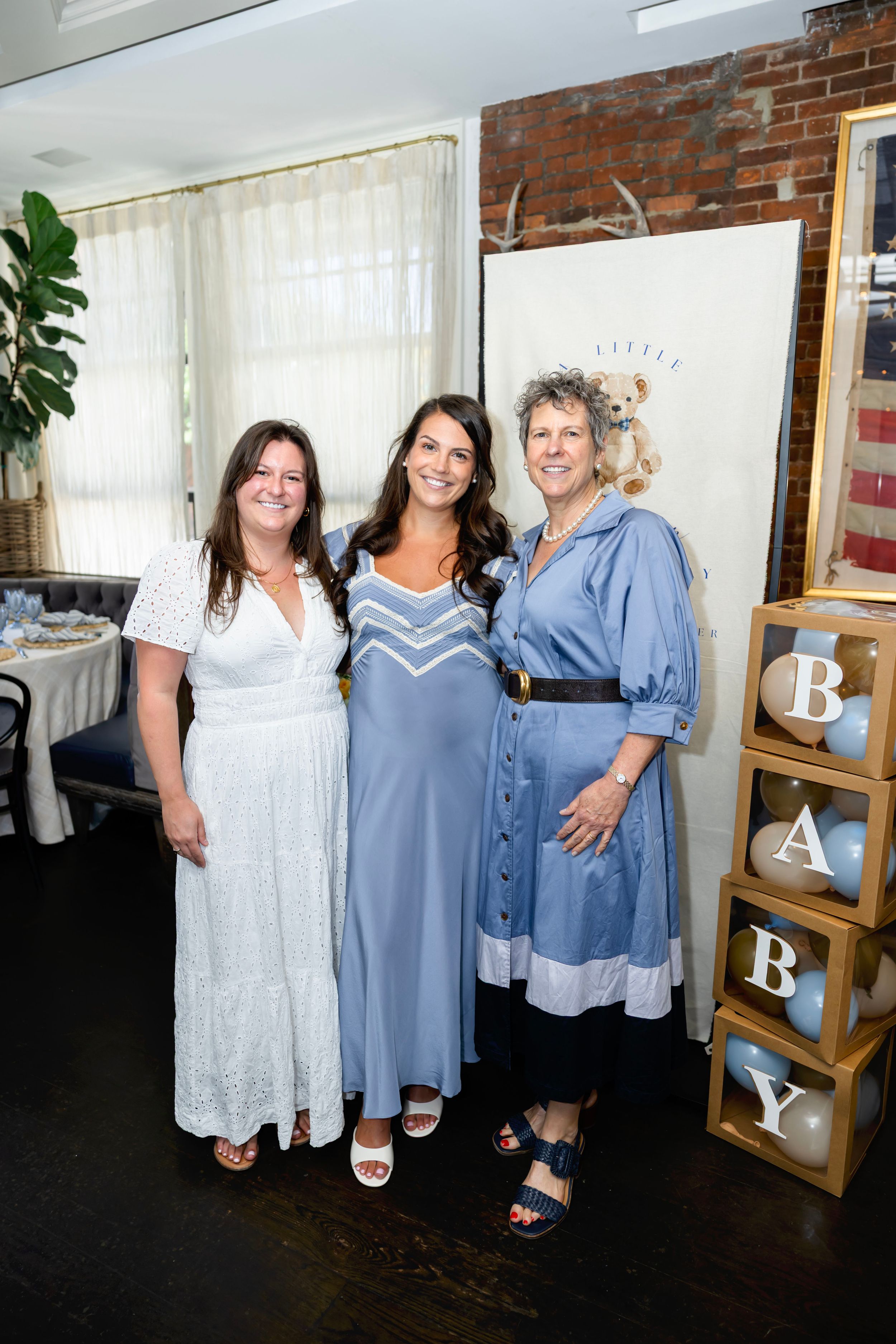 Three women posing together at a baby shower with BABY letter blocks and blue balloon decorations.