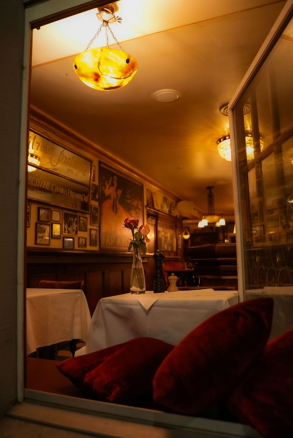 Cozy restaurant interior with warm lighting, white tablecloths, red velvet pillows, and floral centerpiece viewed through window.