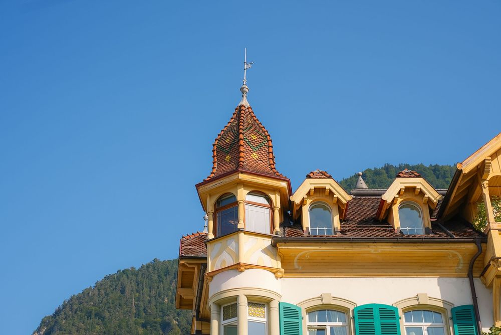 A charming Alpine building with a decorative tower, dormer windows, and green shutters against a clear blue sky.