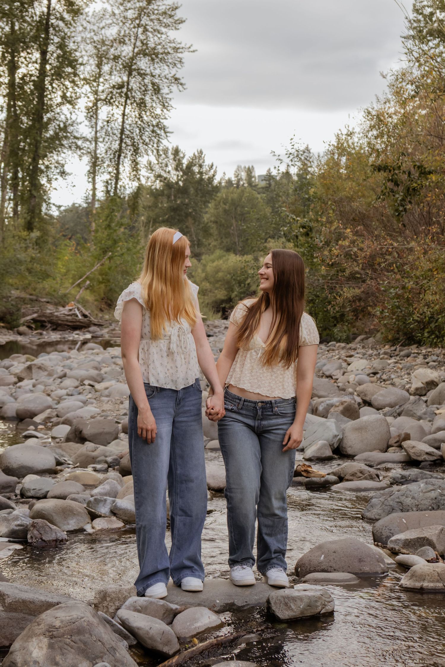 Two teenage girls holding hands and smiling at each other on rocky riverbank surrounded by autumn trees.