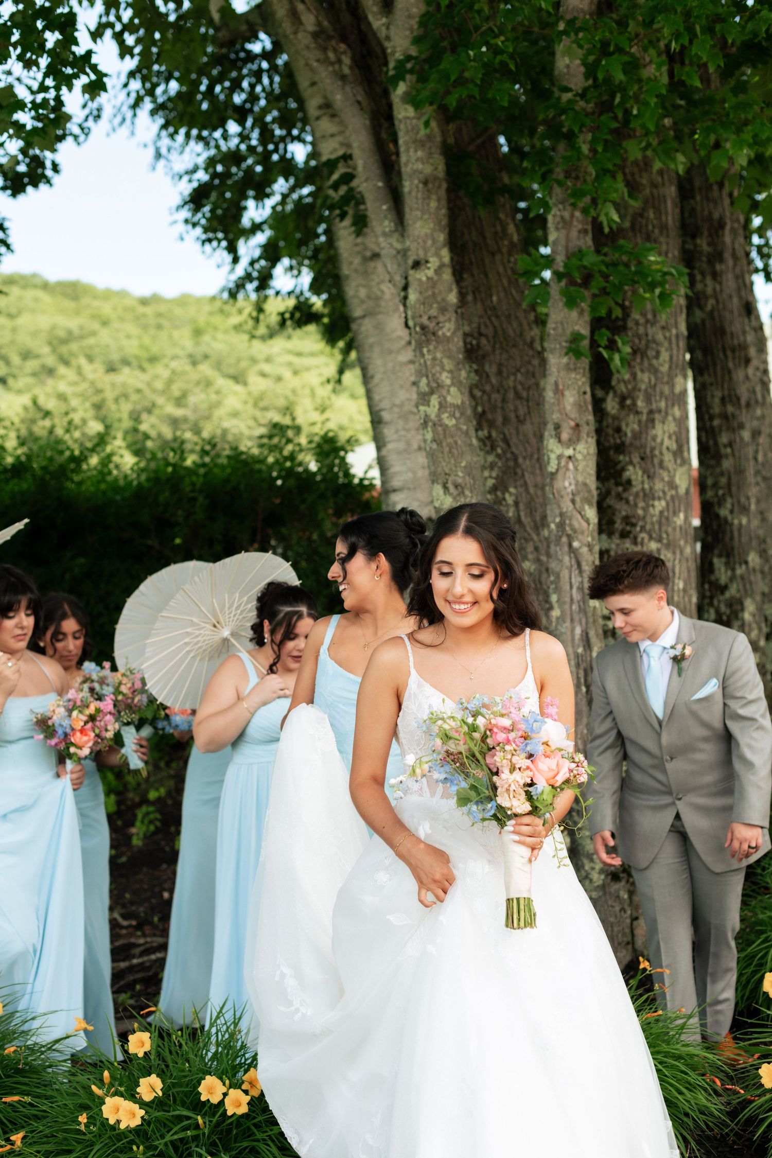 Smiling bride in white gown holds colorful bouquet with bridesmaids in light blue dresses and groomsman outdoors.